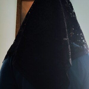 May include: A black lace veil covering a person's head and shoulders. The veil features a delicate, patterned lace design. The person is wearing a teal top. A framed picture is visible in the background.