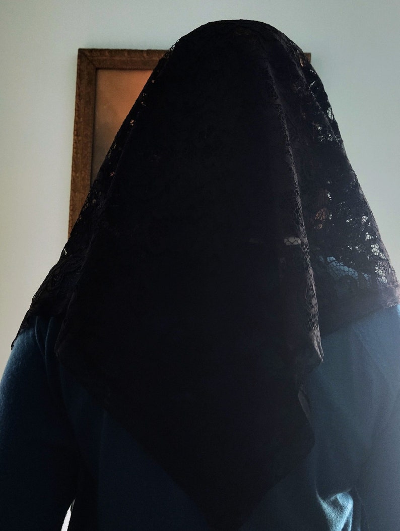 May include: A black lace veil covering a person's head and shoulders. The veil features a delicate, patterned lace design. The person is wearing a teal top. A framed picture is visible in the background.