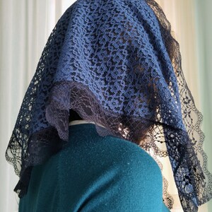 May include: A navy blue lace veil with a scalloped edge. The veil is draped over a person's head and shoulders, with a teal sweater visible underneath. The lace pattern is intricate and detailed.