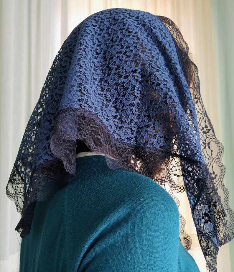May include: A navy blue lace veil with a scalloped edge. The veil is draped over a person's head and shoulders, with a teal sweater visible underneath. The lace pattern is intricate and detailed.