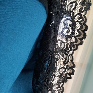 May include: Close-up of a teal garment with black lace trim. The lace features a detailed floral pattern and a scalloped edge. The fabric appears soft and the lace is intricately designed.