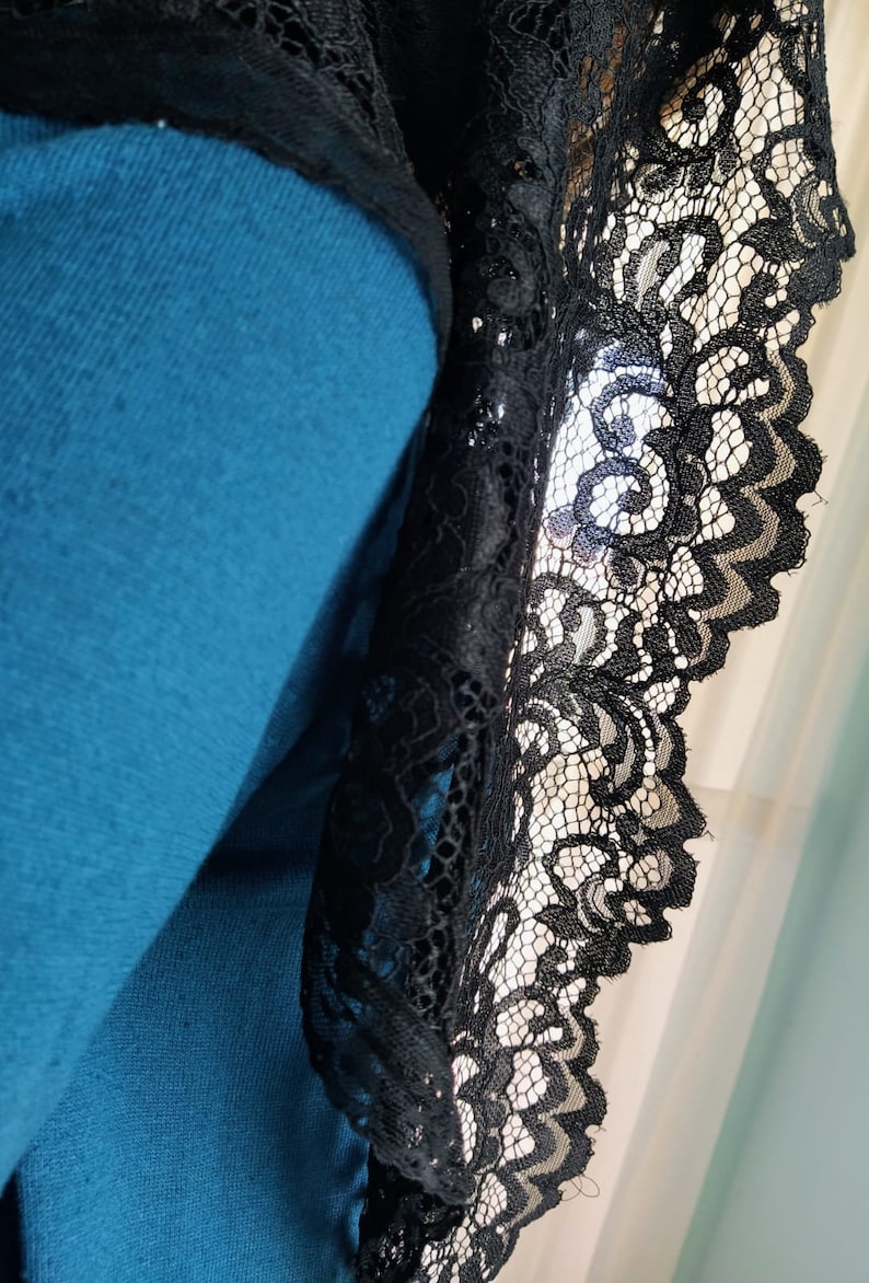 May include: Close-up of a teal garment with black lace trim. The lace features a detailed floral pattern and a scalloped edge. The fabric appears soft and the lace is intricately designed.
