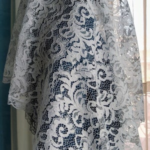 May include: A white lace veil featuring a detailed floral design. The veil is draped, highlighting the delicate openwork pattern. The lace has a scalloped edge, adding elegance. The background includes blue and neutral tones.