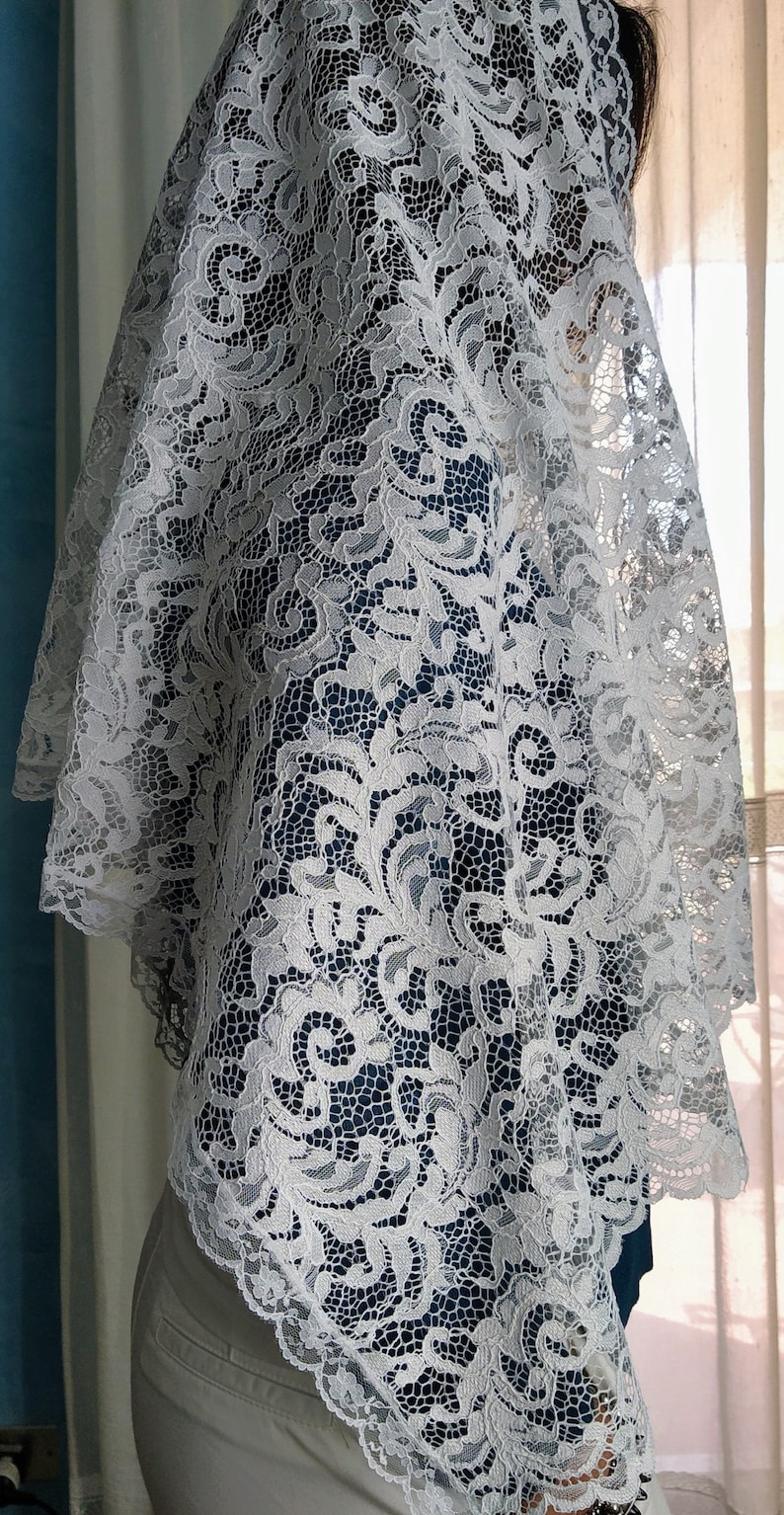 May include: A white lace veil featuring a detailed floral design. The veil is draped, highlighting the delicate openwork pattern. The lace has a scalloped edge, adding elegance. The background includes blue and neutral tones.