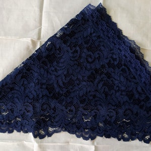 May include: A navy blue lace triangle scarf, featuring detailed floral patterns and a scalloped edge. The scarf is folded and rests on a white fabric background. The lace appears delicate and detailed.