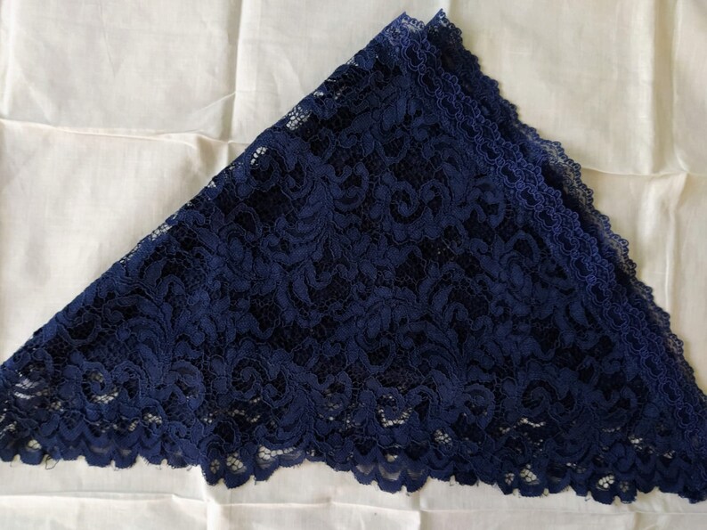 May include: A navy blue lace triangle scarf, featuring detailed floral patterns and a scalloped edge. The scarf is folded and rests on a white fabric background. The lace appears delicate and detailed.