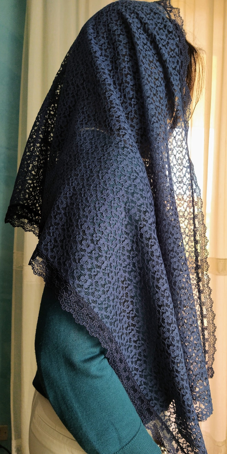 May include: A navy blue lace veil with a scalloped edge. The veil is draped over the head and shoulders, with the intricate floral lace pattern visible. The person is wearing a teal green jumper.