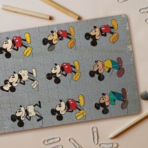 Puede incluir: Rompecabezas vintage de Mickey Mouse con múltiples imágenes de Mickey en varias poses. El rompecabezas está sobre un fondo azul claro con un aspecto desgastado. Lápices, clips y una goma de borrar están esparcidos alrededor del rompecabezas.