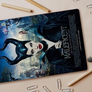 Puede incluir: Un rompecabezas con un póster de la película Maléfica, protagonizada por Angelina Jolie. El póster presenta una combinación de colores azul oscuro y verde con un primer plano de Maléfica, una hada oscura con cuernos, alas y una expresión amenazante. El texto "Disney MALEFICENT" se muestra de forma prominente en el póster.