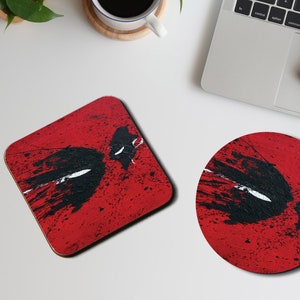 May include: Two coasters featuring a red background with a black and white stylized image of a superhero's mask. The larger coaster is square and the smaller coaster is round.