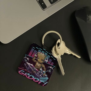 May include: A black square keychain with a colorful image of a cartoon Groot character wearing headphones and holding a record player. The text "I'm Groot!" is written in pink and blue letters on the keychain. The keychain is attached to a silver key ring with two keys.