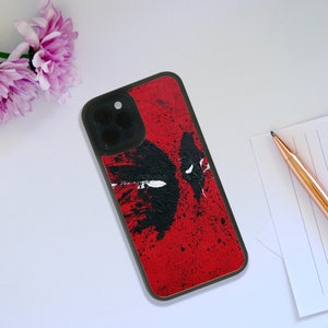 Puede incluir: Una funda de teléfono negra con un diseño de Deadpool rojo y negro. La funda tiene una superficie texturizada y una protuberancia de cámara elevada.