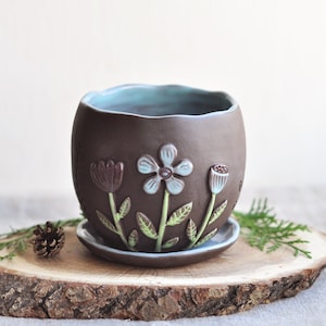 May include: A dark brown ceramic planter with a wavy rim and a matching saucer. The exterior features embossed floral designs with light blue petals, green stems, and leaves. The interior of the planter and the saucer are glazed in a light blue colour.