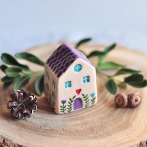 Peut inclure: Figurine de maison en céramique avec un toit violet et un corps beige. La maison a des fenêtres bleues, une porte violette et un cœur rouge. Des fleurs et des feuilles décoratives sont peintes sur la maison. La maison est sur une tranche de bois.