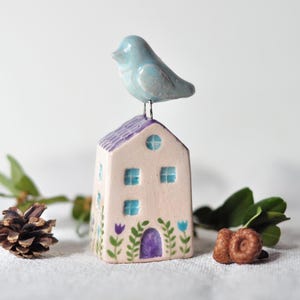 May include: A ceramic house figurine with a light blue bird perched on the purple roof. The house is painted with blue windows, a purple door, and floral accents. Decorative elements include a pine cone and greenery.