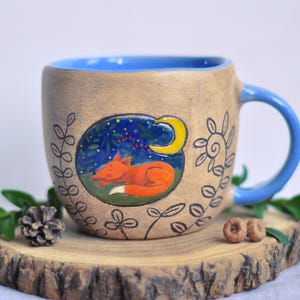 Puede incluir: Una taza de cerámica color canela con interior y asa azules, que presenta una escena pintada a mano de un zorro naranja durmiendo bajo una luna creciente amarilla y un cielo nocturno azul, rodeada de patrones botánicos negros dibujados.