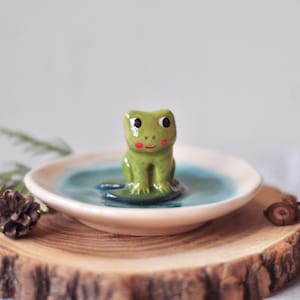 Puede incluir: Una figurita de rana de cerámica sentada en un pequeño plato de cerámica blanco con un centro azul. La rana es verde con mejillas rojas y ojos negros. El plato está sentado en una rebanada de madera.