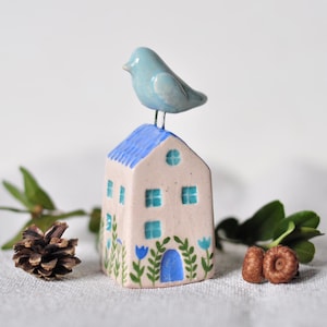 Pode incluir: Uma pequena casa de cerâmica com telhado azul e paredes brancas, decorada com janelas azuis e uma porta azul. Um pássaro azul está sentado no topo do telhado. A casa é cercada por folhas verdes e bolotas marrons.