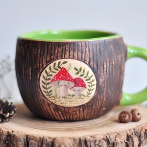 K&ouml;nnte beinhalten: Eine Keramik-Tasse mit einer braunen, strukturierten Au&szlig;enseite, die Baumrinde &auml;hnelt. Der Innenraum und der Griff sind hellgr&uuml;n. Ein kreisf&ouml;rmiges Emblem zeigt rote und wei&szlig;e Pilze. Die Tasse steht auf einer Holzscheibe.