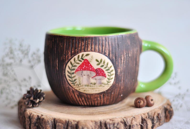 K&ouml;nnte beinhalten: Eine Keramik-Tasse mit einer braunen, strukturierten Au&szlig;enseite, die Baumrinde &auml;hnelt. Der Innenraum und der Griff sind hellgr&uuml;n. Ein kreisf&ouml;rmiges Emblem zeigt rote und wei&szlig;e Pilze. Die Tasse steht auf einer Holzscheibe.