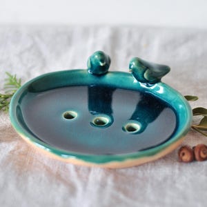 Puede incluir: Un jabonera de cerámica turquesa con dos pequeñas figuras de pájaros posadas en el borde. El plato tiene tres agujeros de drenaje y un borde de color beige claro. Elementos decorativos: piña y ramitas verdes.