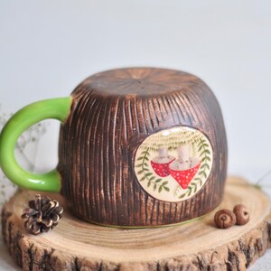 K&ouml;nnte beinhalten: Eine Keramik-Tasse mit brauner, strukturierter Au&szlig;enseite, die Baumrinde &auml;hnelt. Die Tasse hat einen hellgr&uuml;nen Henkel und ein rundes Design mit zwei roten Pilz-Illustrationen. Die Tasse steht auf einer Holzscheibe.