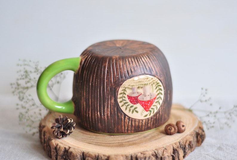K&ouml;nnte beinhalten: Eine Keramik-Tasse mit brauner, strukturierter Au&szlig;enseite, die Baumrinde &auml;hnelt. Die Tasse hat einen hellgr&uuml;nen Henkel und ein rundes Design mit zwei roten Pilz-Illustrationen. Die Tasse steht auf einer Holzscheibe.