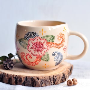 Pode incluir: Uma caneca de cerâmica branca com um design floral colorido. A caneca tem uma alça e está apoiada em um descanso de madeira.