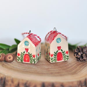 Pode incluir: Dois pequenos ornamentos de cerâmica em forma de casa com paredes cor de creme, telhados vermelhos e portas vermelhas. Cada casa apresenta folhagem verde pintada à mão, tulipas vermelhas e um coração vermelho acima da porta. Uma janela circular azul está na frente e um cordão vermelho e branco está preso ao topo para pendurar.