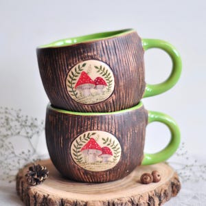 K&ouml;nnte beinhalten: Zwei gestapelte Keramikbecher mit brauner, strukturierter Au&szlig;enseite und gr&uuml;nem Innenraum und Griff. Jeder Becher hat ein kreisf&ouml;rmiges Design mit roten Pilzillustrationen. Die Becher stehen auf einer Holzscheibe.