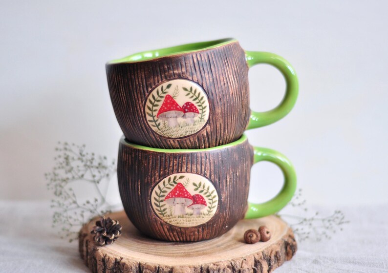 K&ouml;nnte beinhalten: Zwei gestapelte Keramikbecher mit brauner, strukturierter Au&szlig;enseite und gr&uuml;nem Innenraum und Griff. Jeder Becher hat ein kreisf&ouml;rmiges Design mit roten Pilzillustrationen. Die Becher stehen auf einer Holzscheibe.
