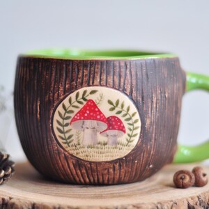 K&ouml;nnte beinhalten: Eine handgefertigte Keramiktasse mit einer braunen, strukturierten Au&szlig;enseite, die Baumrinde &auml;hnelt. Der Innenraum und der Griff sind gr&uuml;n. Ein kreisf&ouml;rmiges Emblem zeigt rot gekappte Pilze. Die Tasse wird auf einer Holzscheibe pr&auml;sentiert.