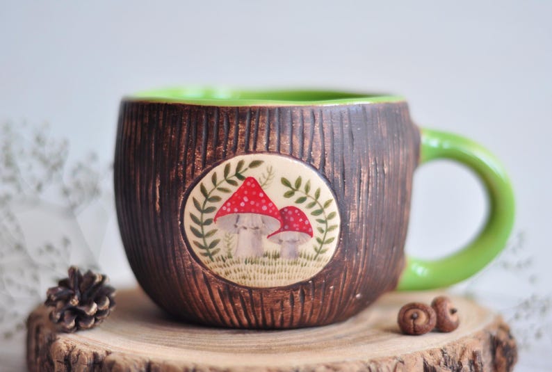K&ouml;nnte beinhalten: Eine handgefertigte Keramiktasse mit einer braunen, strukturierten Au&szlig;enseite, die Baumrinde &auml;hnelt. Der Innenraum und der Griff sind gr&uuml;n. Ein kreisf&ouml;rmiges Emblem zeigt rot gekappte Pilze. Die Tasse wird auf einer Holzscheibe pr&auml;sentiert.