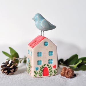 May include: A small ceramic house with a red roof and blue windows. A blue bird sits on top of the house. The house is decorated with flowers and green leaves.