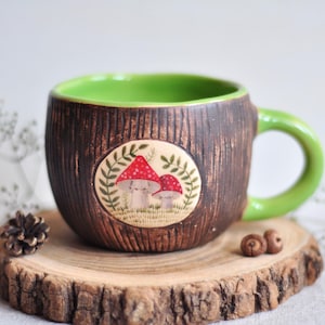 K&ouml;nnte beinhalten: Eine Keramik-Tasse mit brauner, strukturierter Au&szlig;enseite und hellgr&uuml;nem Innenraum und Henkel. Die Tasse hat ein kreisf&ouml;rmiges Design mit zwei rot gekappten Pilzen. Die Tasse wird auf einer Holzscheibe pr&auml;sentiert.