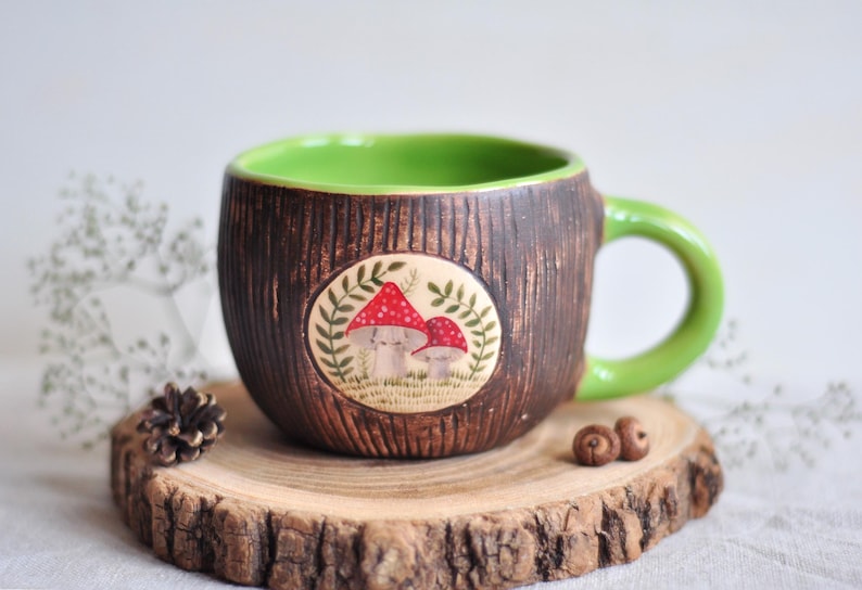 K&ouml;nnte beinhalten: Eine Keramik-Tasse mit brauner, strukturierter Au&szlig;enseite und hellgr&uuml;nem Innenraum und Henkel. Die Tasse hat ein kreisf&ouml;rmiges Design mit zwei rot gekappten Pilzen. Die Tasse wird auf einer Holzscheibe pr&auml;sentiert.