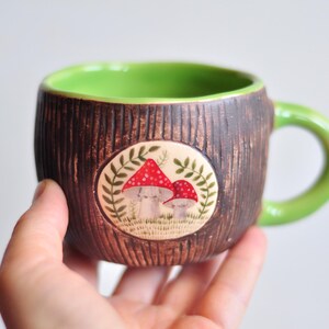 K&ouml;nnte beinhalten: Eine Keramik-Tasse mit brauner, strukturierter Au&szlig;enseite und hellgr&uuml;nem Innenraum und Henkel. Die Tasse hat ein kreisf&ouml;rmiges Design mit zwei rot gekappten Pilzen und gr&uuml;nem Laub. Die Tasse wird in einer Hand gehalten.