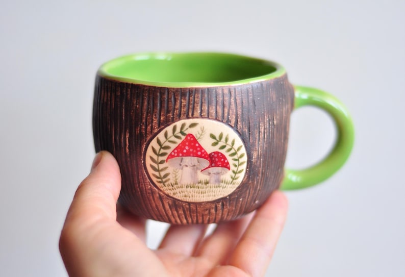 K&ouml;nnte beinhalten: Eine Keramik-Tasse mit brauner, strukturierter Au&szlig;enseite und hellgr&uuml;nem Innenraum und Henkel. Die Tasse hat ein kreisf&ouml;rmiges Design mit zwei rot gekappten Pilzen und gr&uuml;nem Laub. Die Tasse wird in einer Hand gehalten.