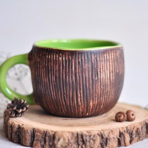 K&ouml;nnte beinhalten: Eine handgefertigte Keramik-Tasse mit einer strukturierten, braunen Au&szlig;enseite und einem hellgr&uuml;nen Innenraum und Griff. Die Tasse steht auf einer Holzscheibe mit einem kleinen Tannenzapfen und zwei Eicheln. Ein einzigartiges, rustikales Design.