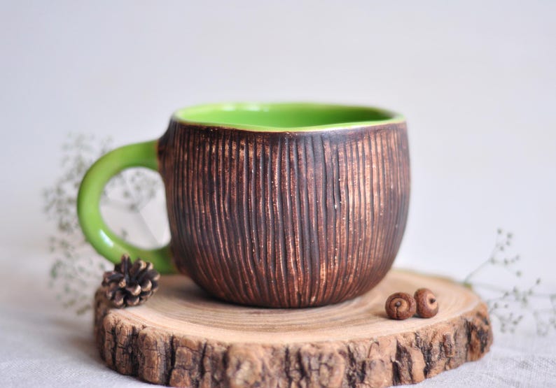 K&ouml;nnte beinhalten: Eine handgefertigte Keramik-Tasse mit einer strukturierten, braunen Au&szlig;enseite und einem hellgr&uuml;nen Innenraum und Griff. Die Tasse steht auf einer Holzscheibe mit einem kleinen Tannenzapfen und zwei Eicheln. Ein einzigartiges, rustikales Design.