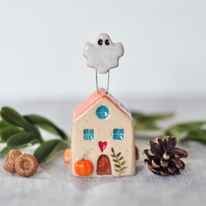 Puede incluir: Adorno de cerámica con forma de casa con techo naranja, ventanas azules y un corazón rojo. Una pequeña figura de fantasma blanco está unida a la parte superior. La casa tiene detalles de calabaza y hojas. También hay una piña y bellotas.