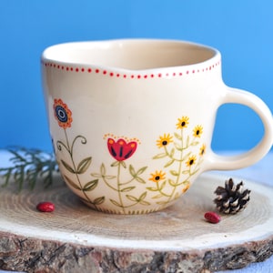 Pode incluir: Caneca de cerâmica creme com um design floral pintado à mão. A caneca apresenta flores vermelhas, amarelas e azuis, com caules e folhas verdes. A borda é pontilhada com vermelho. A caneca está em uma fatia de madeira.