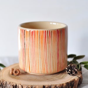 Op de afbeelding: Cilindrische keramische plantenpot met verticale strepen in rood, oranje en bruin. De pot staat op een houten schijf met een dennenappel en groen. De binnenkant van de pot is lichtbeige.