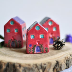 May include: Three miniature ceramic houses with red walls and purple or gray roofs are displayed on a wooden slice. Each house has turquoise windows and a door with floral accents. A pinecone and greenery are also present.