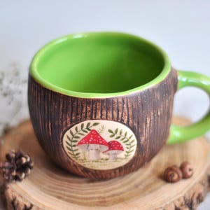 K&ouml;nnte beinhalten: Eine Keramik-Tasse mit braunem, strukturiertem &Auml;u&szlig;eren und einem hellgr&uuml;nen Innenraum und Griff. Die Tasse hat ein ovales Design mit roten und wei&szlig;en Pilzen. Die Tasse steht auf einem Holzuntersetzer.