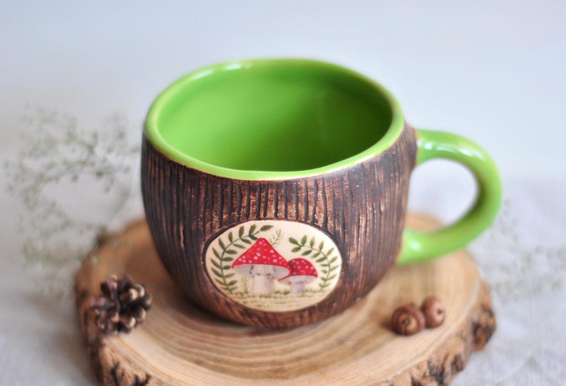 K&ouml;nnte beinhalten: Eine Keramik-Tasse mit braunem, strukturiertem &Auml;u&szlig;eren und einem hellgr&uuml;nen Innenraum und Griff. Die Tasse hat ein ovales Design mit roten und wei&szlig;en Pilzen. Die Tasse steht auf einem Holzuntersetzer.