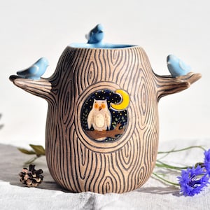 May include: A ceramic tree-shaped vase with a light blue interior and three light blue birds perched on the branches. The vase features a circular design with an owl, moon, and stars.