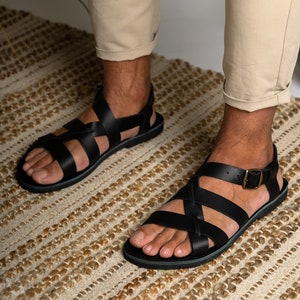 May include: Black leather sandals featuring a multi-strap design that crosses over the top of the foot. The sandals include an adjustable ankle strap with a metallic buckle closure and a flat, dark sole, providing a secure fit for the wearer.