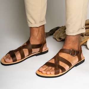 May include: A pair of dark brown leather sandals featuring multiple crisscrossing straps across the top of the foot. The sandals include a side buckle closure and a flat black sole. The design consists of wide leather bands that secure the foot.