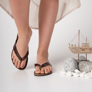 May include: A pair of brown leather thong sandals with a simple design. The sandals are being worn by a person standing on a white surface. A small wooden sailboat is placed on a pile of grey and white stones.